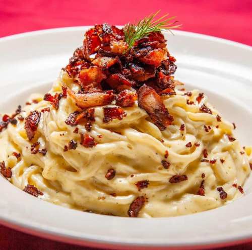 Creamy pasta topped with crispy bacon bits and a sprig of dill, served in a white bowl on a red tablecloth.