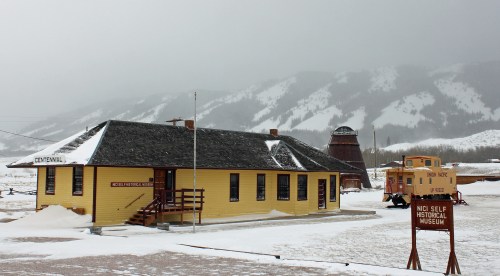 A yellow historical museum building with snow-covered ground, surrounded by mountains and a vintage train car nearby.