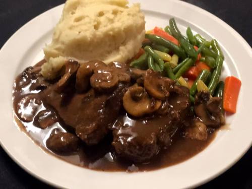A plate of beef with mushroom gravy, mashed potatoes, and a side of green beans and mixed vegetables.