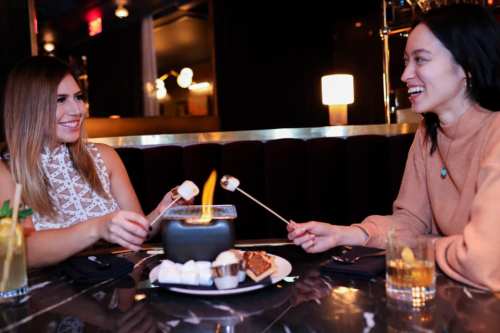 Two women sitting at a table in a restaurant, roasting marshmallows over a small flame, smiling and enjoying their time.