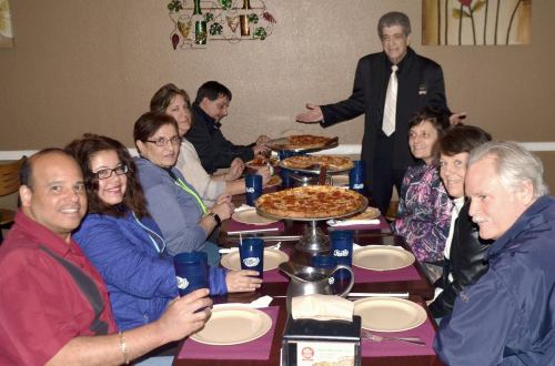 A group of people enjoying a meal at a restaurant, with a large pizza and a waiter presenting the food.