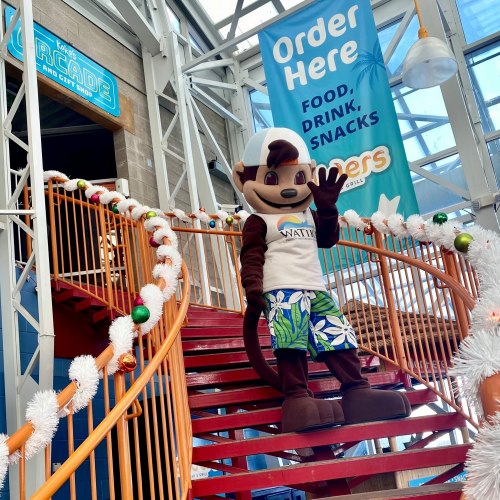 A mascot in a white shirt and colorful shorts waves from a staircase in a festive arcade setting.