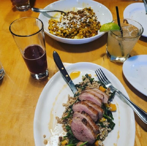 A plate with sliced duck breast on greens and grains, alongside a bowl of pasta and drinks on a wooden table.