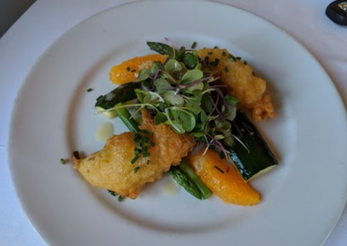 A plate of fried fish with zucchini, asparagus, orange segments, and microgreens, drizzled with a light sauce.