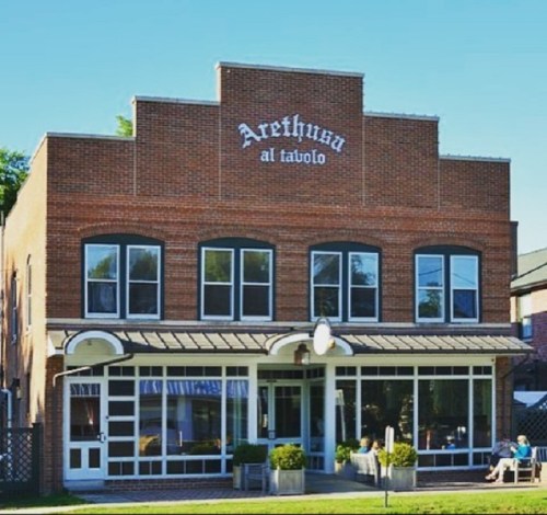 A brick building with large windows and a sign reading "Arethusa al tavolo," set against a clear blue sky.