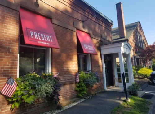 Exterior of a brick building with red awnings labeled "PRESENT company," featuring American flags and greenery.