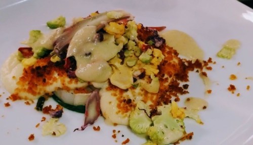 A plated dish featuring crispy breaded fish topped with sauce, vegetables, and crushed nuts on a white background.