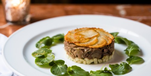 A gourmet dish featuring layered meat topped with crispy potatoes, surrounded by fresh greens on a white plate.