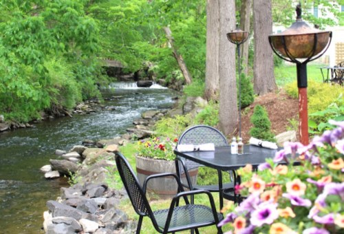 A serene outdoor dining area by a flowing stream, surrounded by greenery and colorful flowers.