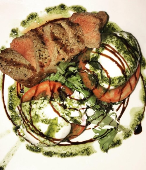 A plate featuring grilled steak, poached eggs, tomatoes, and drizzled with pesto and balsamic reduction.