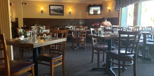 A cozy restaurant interior with empty tables set for dining and a patron seated in the background.