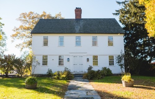 A white two-story house with a gray roof, surrounded by greenery and a stone pathway leading to the front door.