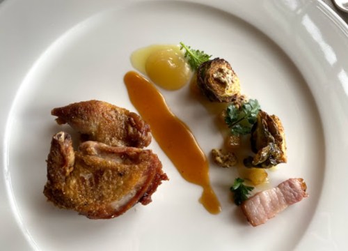 A plated dish featuring crispy chicken, sauce, Brussels sprouts, and a slice of bacon, garnished with herbs.
