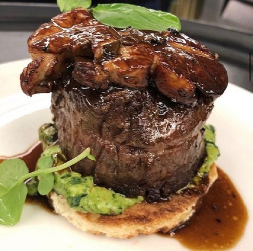 A gourmet dish featuring a tender beef fillet topped with sautéed mushrooms, served on a bed of green sauce and toast.