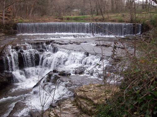 A serene waterfall cascading over rocky ledges, surrounded by trees and greenery in a tranquil natural setting.