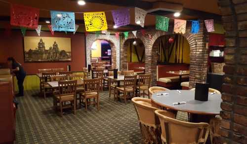 A cozy restaurant interior with wooden tables, colorful papel picado decorations, and a brick archway.