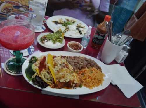 A colorful plate of tacos, rice, beans, and guacamole, with drinks and additional dishes in the background.