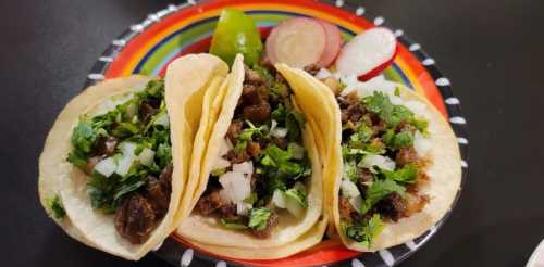 Three tacos filled with meat, topped with onions and cilantro, served with lime and radishes on a colorful plate.