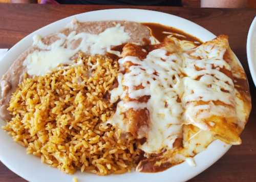 A plate of enchiladas topped with cheese, served with rice and refried beans on the side.