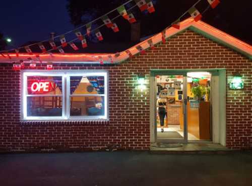 A brick building with an "OPEN" sign, colorful lights, and Mexican flags, creating a welcoming atmosphere at night.