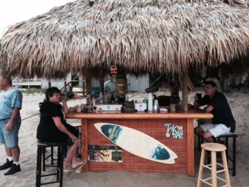A beach bar with a thatched roof, featuring patrons sitting at the counter and a surfboard decoration.