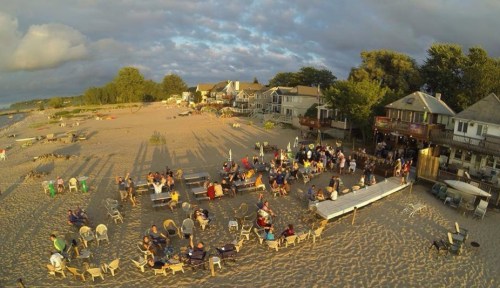 A beach scene with people gathered around tables, enjoying the outdoors, with houses and trees in the background.