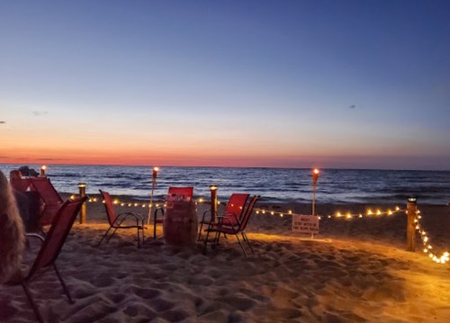 A serene beach at sunset, with chairs, lanterns, and gentle waves under a colorful sky.