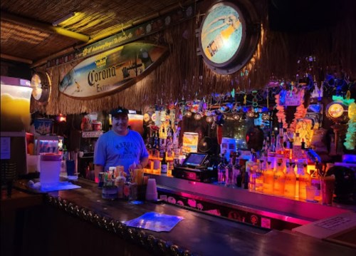 A vibrant bar scene with colorful lights, bottles, and a bartender in a casual setting. Tropical decor is visible.