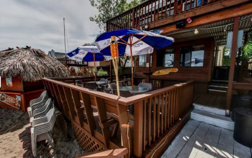 A beachside bar with wooden decking, umbrellas, and seating, surrounded by sand and tropical decor.