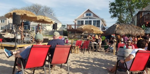 A lively beach scene with people enjoying music, seated in chairs under umbrellas, with a band performing on stage.