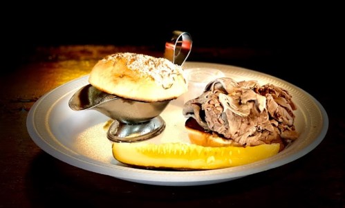 A plate with a bun, sliced roast beef, a pickle, and a small cup of gravy.