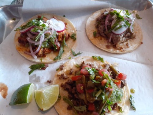 Three tacos on a tray: one with chicken, one with beef, and one with mixed toppings, garnished with cilantro and lime.