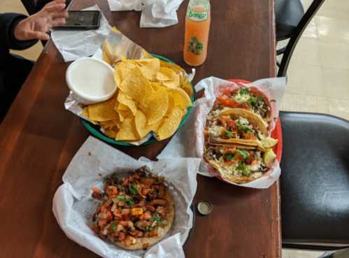 A table with a plate of tacos, a bowl of salsa, tortilla chips, and a bottle of soda.