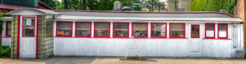 A vintage diner with a curved roof, red trim, and large windows, set on a sidewalk surrounded by greenery.