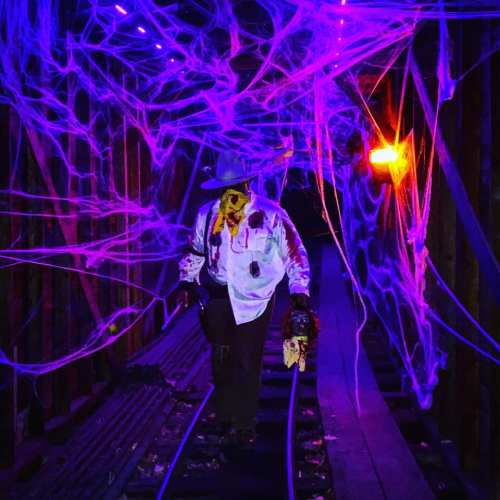 A spooky figure in a white shirt walks through a dark, web-filled tunnel illuminated by purple and orange lights.