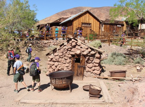 A group of people walks past a stone structure and a large pot, with wooden buildings and hills in the background.