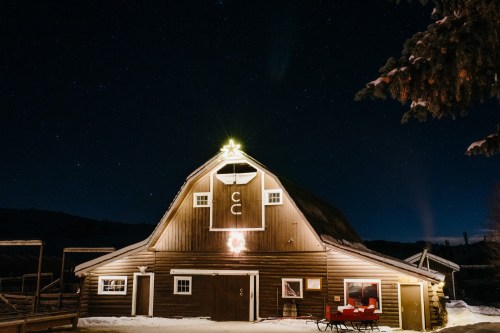 A rustic barn illuminated at night, adorned with a star and wreath, surrounded by snow and a starry sky.