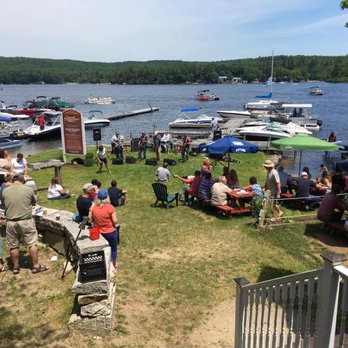 A lively lakeside gathering with people enjoying music, boats docked, and a sunny day by the water.