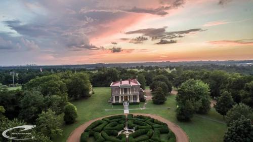 Aerial view of a historic mansion surrounded by lush greenery and a beautifully landscaped garden at sunset.