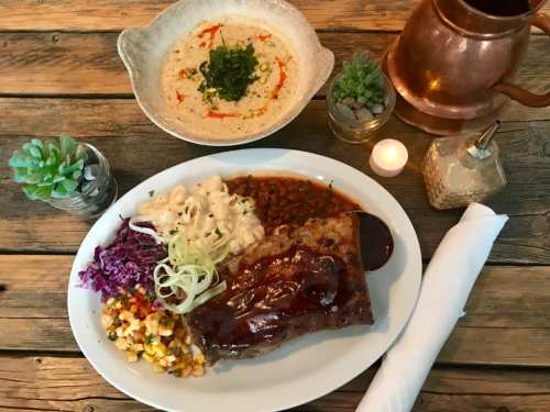 A plate of barbecue meat with sides of coleslaw, corn salad, and creamy pasta, alongside a bowl of soup and decor.