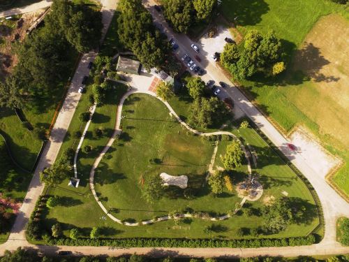 Aerial view of a park featuring winding paths, green lawns, trees, and parked cars nearby.