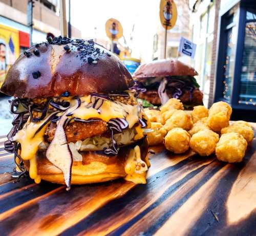 A close-up of a loaded burger with melted cheese, coleslaw, and crispy toppings, alongside golden tater tots on a wooden board.
