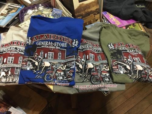Four t-shirts featuring a graphic of the Aladdin General Store, each in different colors: blue, gray, green, and beige.