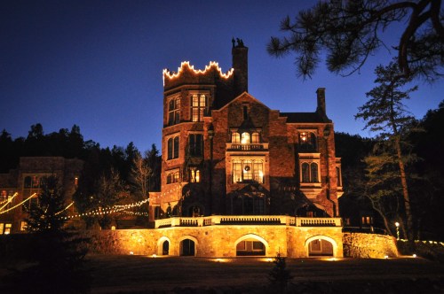 A beautifully lit stone mansion at dusk, surrounded by trees and festive lights.