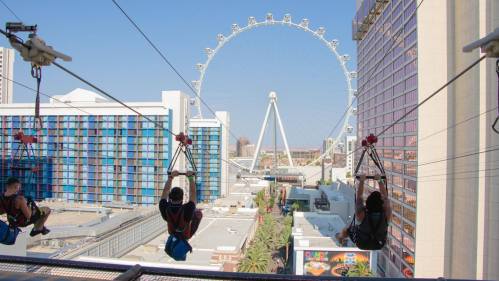 Three people ziplining with a large Ferris wheel in the background against a clear blue sky.