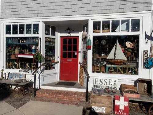 A quaint shop with a red door, showcasing vintage items and a sailboat in the window, near a fried clams sign.