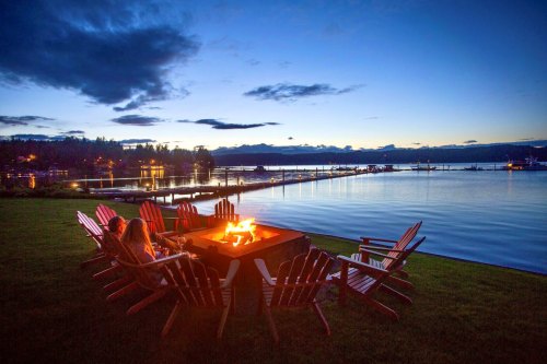 A serene lakeside scene at dusk, featuring a fire pit surrounded by red chairs and calm water reflecting the sky.
