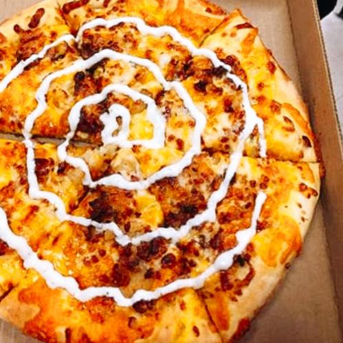 A pizza with a spiral pattern of toppings and white sauce drizzled on top, served in a cardboard box.