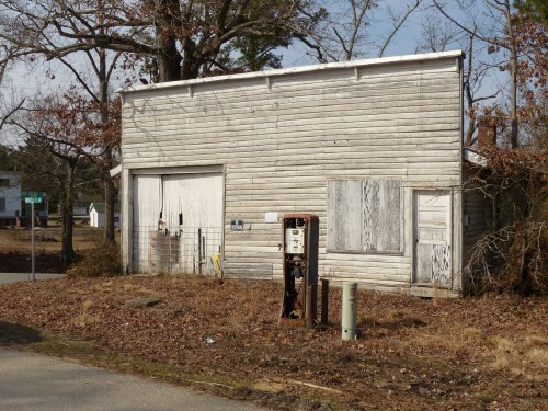 A weathered wooden building with a closed garage door and an old gas pump in front, surrounded by bare trees.