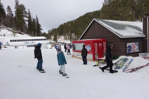 People walking in a snowy area near a ski lodge, with ski equipment and a vending machine visible.